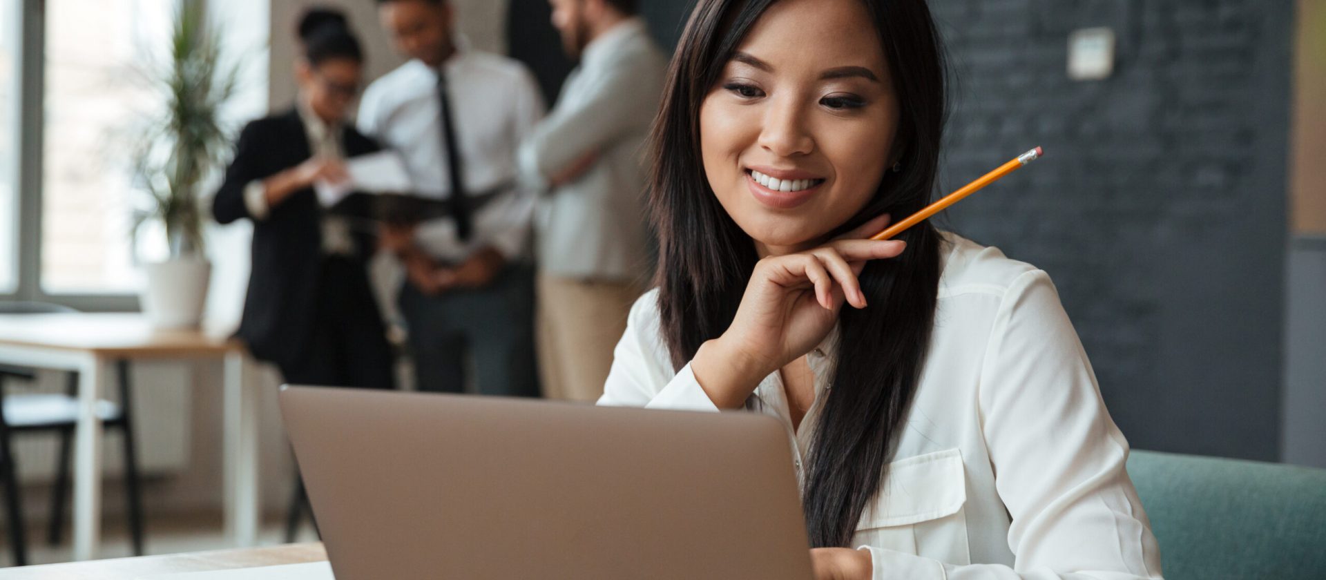 Picture of cheerful young asian business woman sitting indoors using laptop computer. Looking aside.