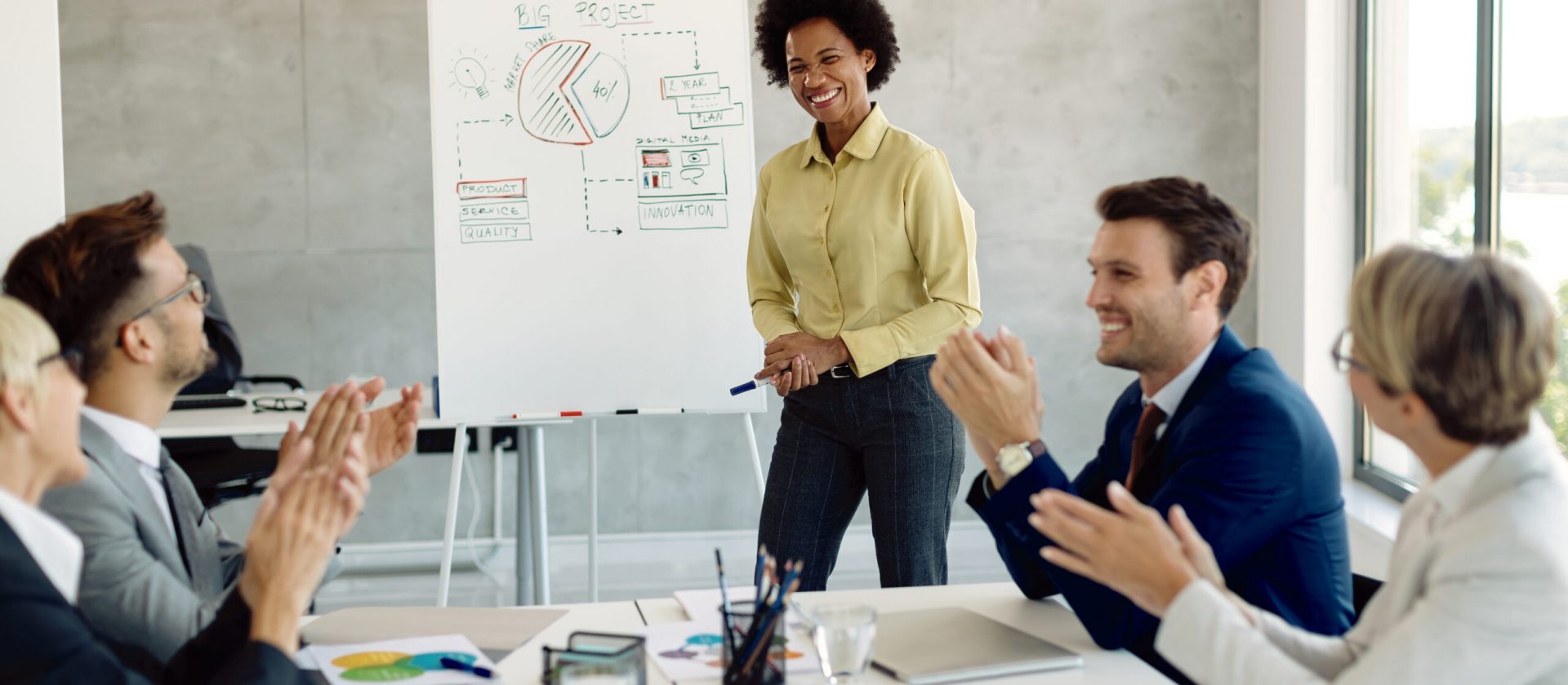 Group of business people applauding to female colleague after successful presentation in the office. Focus is on black businesswoman in front of whiteboard.